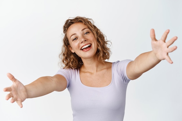 Heureuse fille blonde atteignant un câlin avec un grand sourire amical, attendant des câlins, debout sur fond blanc