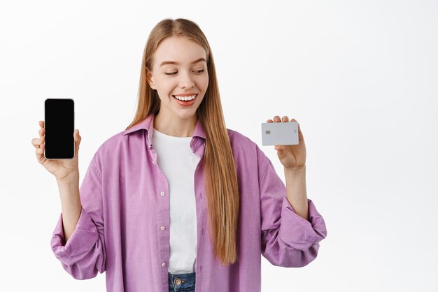 Heureuse fille blonde regardant sa carte de crédit bancaire et montrant l'écran vide du téléphone mobile d'affichage de l'application smartphone souriant heureux debout sur fond blanc