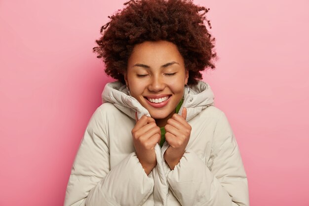 Heureuse fille charmante se sent au chaud dans une veste blanche, sourit largement, garde les yeux fermés, montre des dents parfaites, se dresse sur fond de studio rose