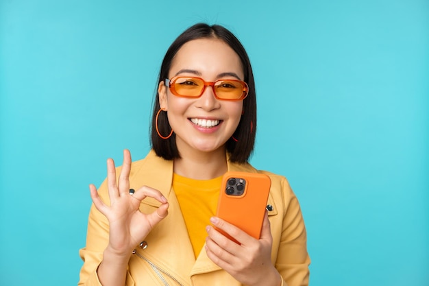 Heureuse fille coréenne à lunettes de soleil montrant un signe correct et tenant un téléphone portable à l'aide d'une application pour smartphone recommandant une application debout sur fond bleu