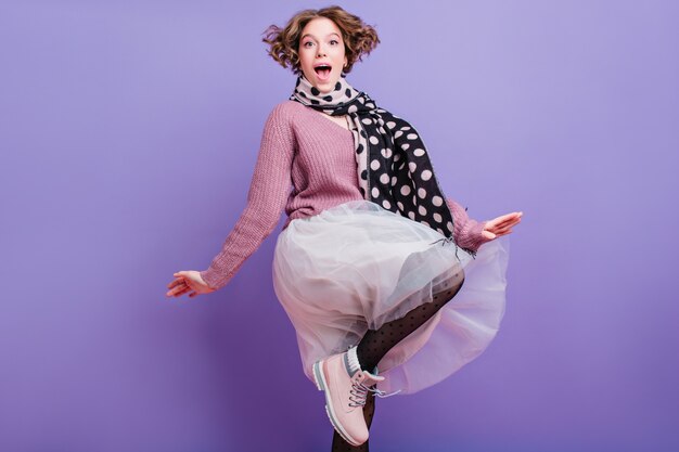 Heureuse fille drôle avec une coiffure frisée courte sautant Photo intérieure d'une jeune femme heureuse en jupe luxuriante et écharpe s'amusant.