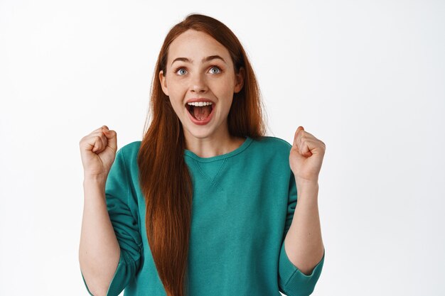 Heureuse fille rousse chanceuse, levant les yeux et chantant, gagnant de l'argent pari, célébrant la victoire, triomphant de joie, enracinant pour l'équipe, debout sur blanc