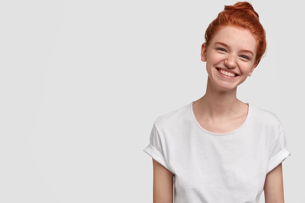 Heureuse fille souriante et insouciante aux taches de rousseur incline la tête, a un sourire à pleines dents, une peau tachetée de rousseur, vêtue de vêtements décontractés, des modèles contre un mur blanc avec espace de copie pour votre publicité ou texte promotionnel