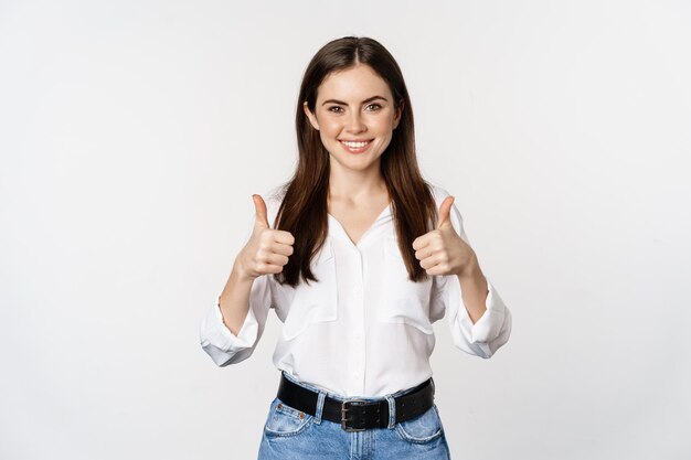 Heureuse fille souriante montrant les pouces vers le haut, étudiante avec des commentaires satisfaits, recommandant qch, debout sur fond blanc