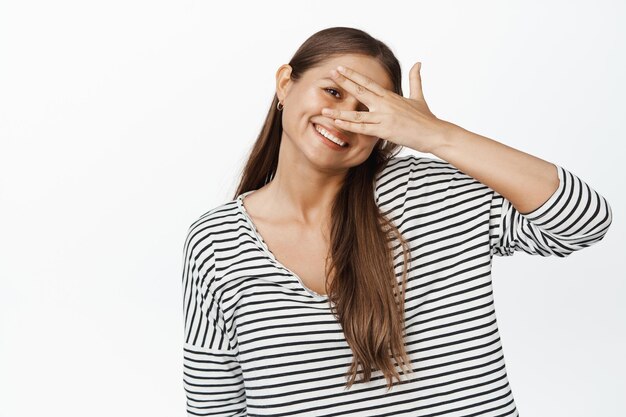 Heureuse fille souriante regardant à travers les doigts, tenant la main contre les yeux, fond blanc