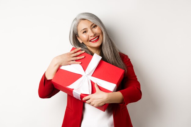 Heureuse grand-mère asiatique étreignant la boîte-cadeau rouge et souriant reconnaissant, remerciant pour le présent, debout sur fond blanc.