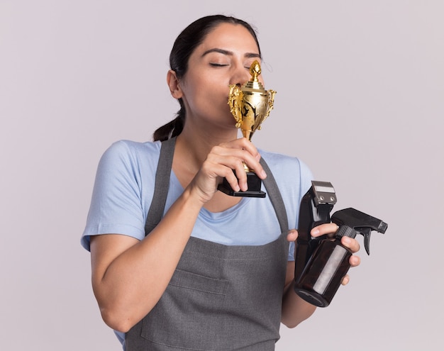 Heureuse jeune belle femme coiffeur en tablier tenant la machine de tondeuse trophée d'or avec vaporisateur embrassant son trophée debout sur un mur blanc