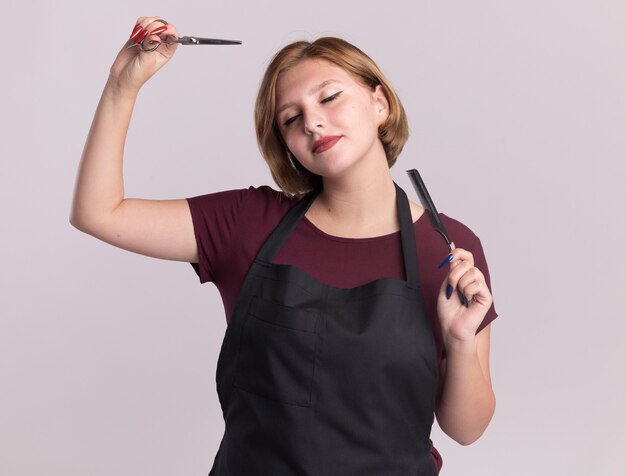 Heureuse jeune belle femme coiffeur en tablier tenant un peigne à cheveux et des ciseaux aux yeux fermés souriant debout sur un mur blanc