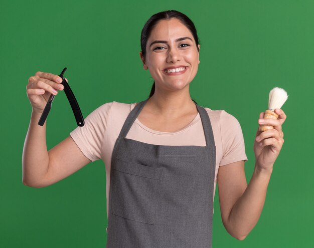 Heureuse jeune belle femme coiffeur en tablier tenant le rasoir et le blaireau à l'avant souriant joyeusement debout sur le mur vert