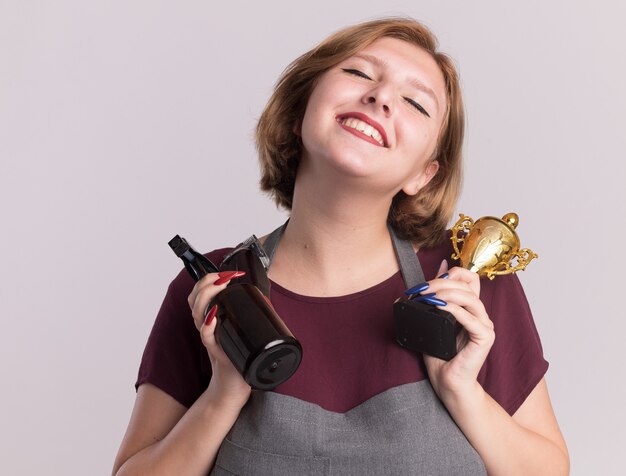 Heureuse jeune belle femme coiffeur en tablier tenant le trophée d'or et vaporisateur avec tondeuse souriant avec les yeux fermés debout sur un mur blanc