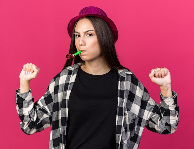 Heureuse jeune belle femme portant un chapeau de fête soufflant un sifflet de fête montrant un geste oui isolé sur un mur rose