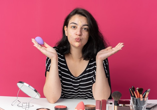 Photo gratuite heureuse jeune belle fille assise à table avec des outils de maquillage appliquant une crème tonifiante tenant une éponge écartant les mains montrant un geste de baiser isolé sur un mur rose