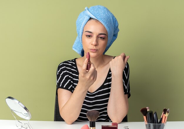 Heureuse jeune belle fille assise à table avec des outils de maquillage enveloppés de cheveux dans une serviette tenant et regardant le rouge à lèvres isolé sur un mur vert olive