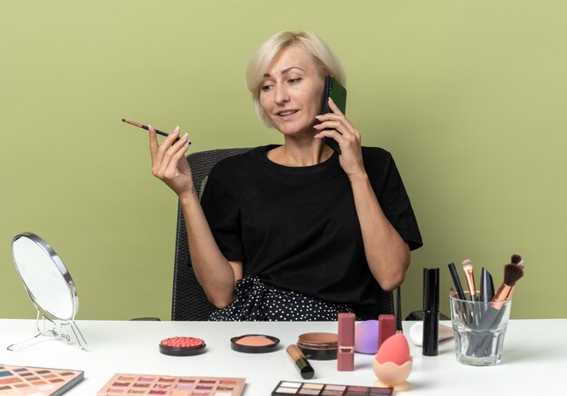 Heureuse jeune belle fille assise à table avec des outils de maquillage parle au téléphone tenant un pinceau de maquillage isolé sur un mur vert olive