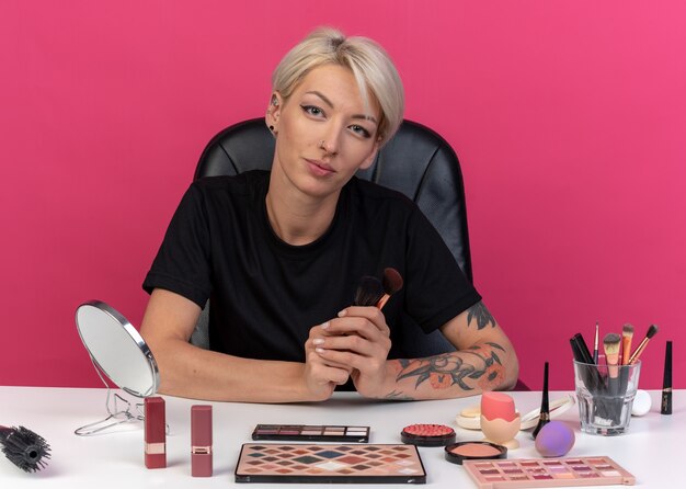 Heureuse jeune belle fille assise à table avec des outils de maquillage tenant des pinceaux à poudre isolés sur un mur rose