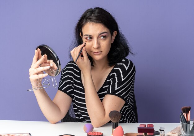 Heureuse jeune belle fille assise à table avec des outils de maquillage tenant et regardant un miroir appliquant un fard à paupières avec un pinceau de maquillage isolé sur un mur bleu