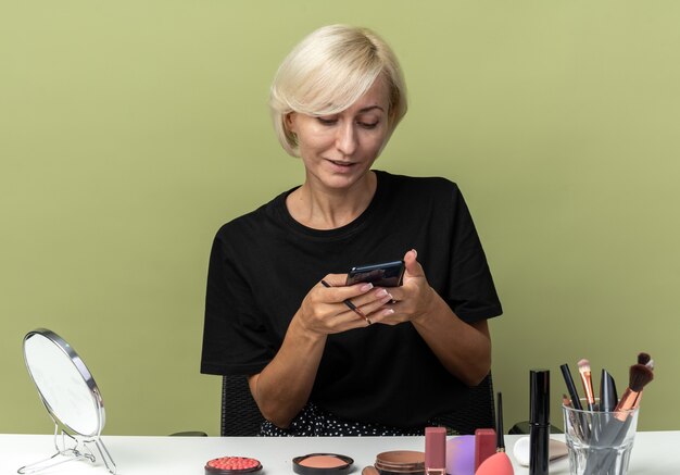 Heureuse jeune belle fille assise à table avec des outils de maquillage tenant et regardant le téléphone dans sa main isolée sur un mur vert olive