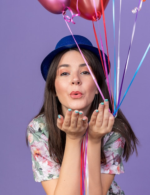 Heureuse jeune belle fille portant un chapeau de fête tenant des ballons montrant un geste de baiser isolé sur un mur bleu