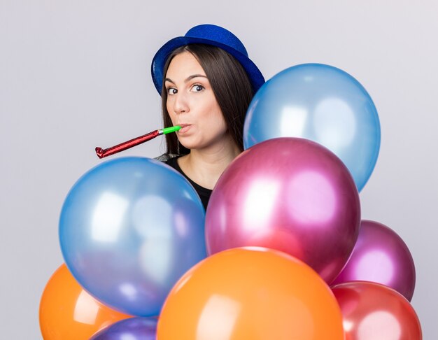 Heureuse jeune belle fille portant un chapeau de fête tenant des ballons soufflant un sifflet de fête