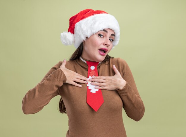 heureuse jeune belle fille portant un chapeau de noël et une cravate mettant les mains sur elle-même isolée sur un mur vert olive