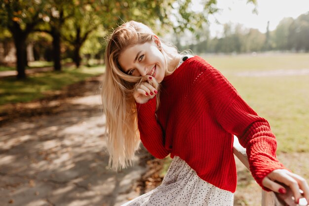 Heureuse jeune blonde en pull rouge à la mode et jupe blanche souriant dans le parc de l'automne. Fille élégante avec un maquillage naturel posant en plein air.