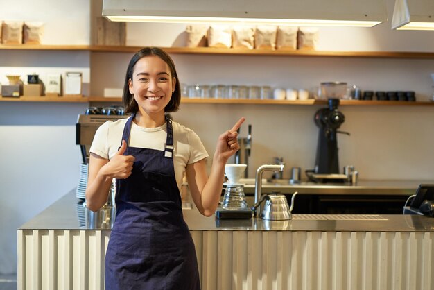 Heureuse jeune employée de personnel barista asiatique en tablier montrant les pouces vers le haut et pointant vers l'espace de copie