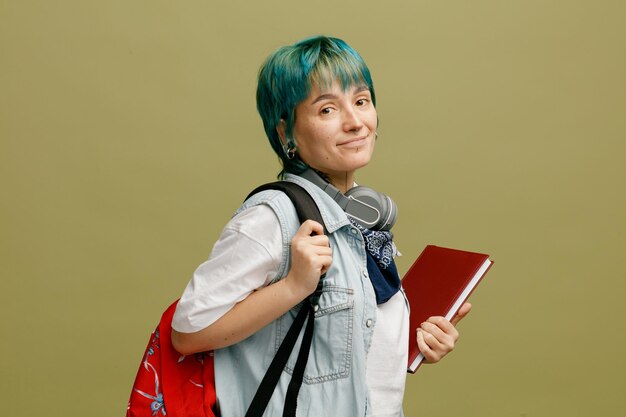 Heureuse jeune étudiante portant des écouteurs et un bandana sur le cou et le sac à dos debout dans la vue de profil saisissant le sac à dos tenant le carnet de notes regardant la caméra isolée sur fond vert olive