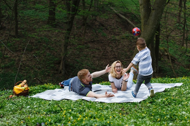 Heureuse jeune famille joue avec un ballon sur le plaid pendant un pique-nique dans le parc