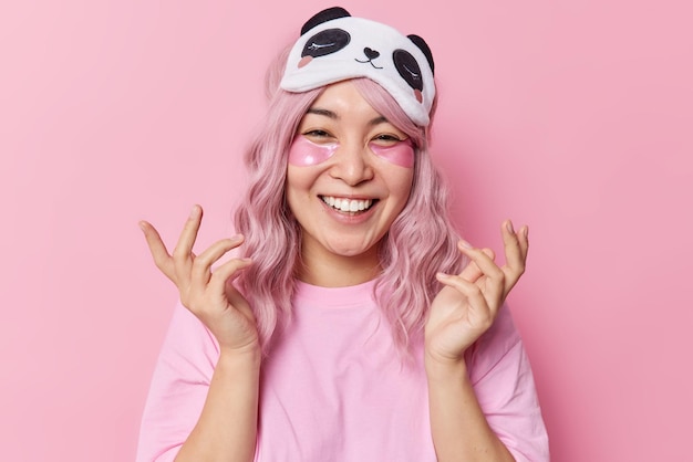 Heureuse jeune femme asiatique aux cheveux roses teints sourit à pleines dents applique des patchs d'hydrogel sous les yeux garde les mains levées aime les procédures de beauté après avoir dormi porte les yeux bandés sur le front et le t-shirt