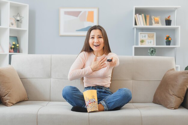 Heureuse jeune femme asiatique en tenue décontractée assise sur un canapé à l'intérieur de la maison avec un seau de pop-corn tenant à distance en regardant la télévision être joyeuse en riant