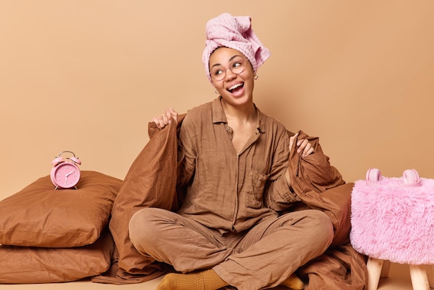 Heureuse jeune femme assise en posture de lotus sur un lit moelleux porte une serviette enveloppée de pyjama sur la tête rit joyeusement se sent détendue après avoir dormi recouvert d'une couverture entourée d'oreillers et d'un réveil