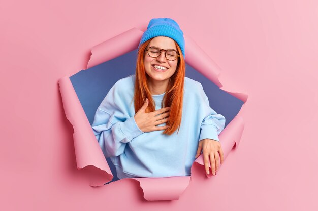 Heureuse jeune femme au gingembre sincère rit fort sourit largement et ne peut pas arrêter de rire garde la main sur la poitrine vêtue d'une tenue bleue élégante perce le mur de papier rose entend une anecdote drôle