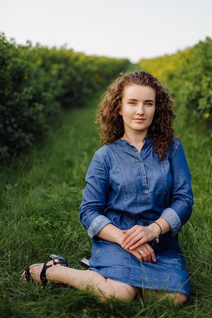 Heureuse jeune femme aux cheveux bouclés bruns, vêtue d'une robe, posant à l'extérieur dans un jardin