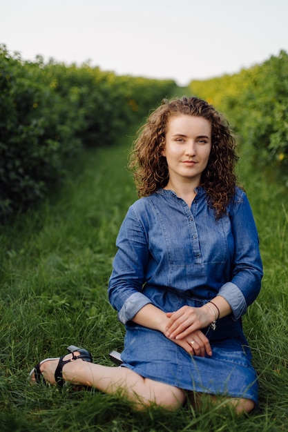 Heureuse jeune femme aux cheveux bouclés bruns, vêtue d'une robe, posant à l'extérieur dans un jardin