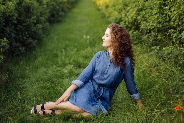 Heureuse jeune femme aux cheveux bouclés bruns, vêtue d'une robe, posant à l'extérieur dans un jardin