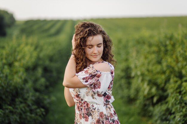 Heureuse jeune femme aux cheveux bouclés bruns, vêtue d'une robe, posant à l'extérieur dans un jardin