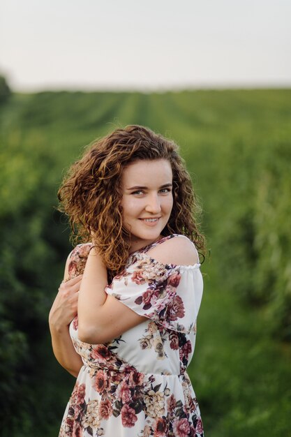 Heureuse jeune femme aux cheveux bouclés bruns, vêtue d'une robe, posant à l'extérieur dans un jardin