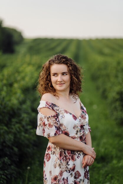 Heureuse jeune femme aux cheveux bouclés bruns, vêtue d'une robe, posant à l'extérieur dans un jardin