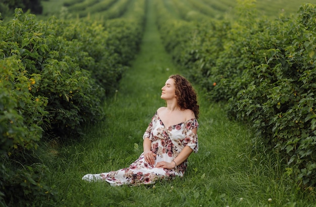 Heureuse jeune femme aux cheveux bouclés bruns, vêtue d'une robe, posant à l'extérieur dans un jardin