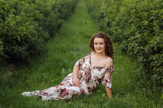 Heureuse jeune femme aux cheveux bouclés bruns, vêtue d'une robe, posant à l'extérieur dans un jardin