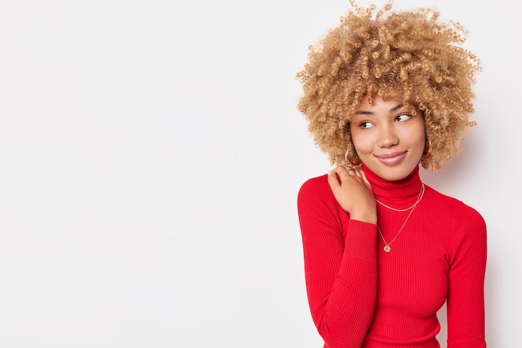 Photo gratuit heureuse jeune femme aux cheveux bouclés garde la main sur l'épaule porte un col roulé rouge confortable pense à quelque chose d'agréable isolé sur fond blanc avec un espace de copie pour votre publicité