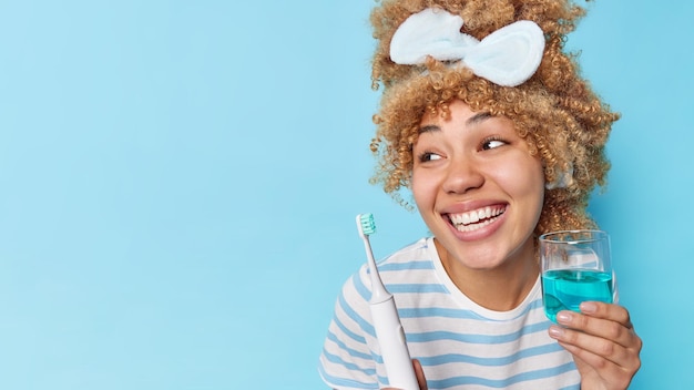 Heureuse jeune femme aux cheveux bouclés tient un verre de rince-bouche frais et une brosse à dents électrique subit des procédures d'hygiène regarde loin isolé sur fond bleu copiez l'espace pour placer votre publicité