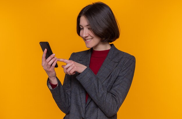 Heureuse jeune femme aux cheveux courts portant une veste grise tenant un smartphone en tapant divers messages