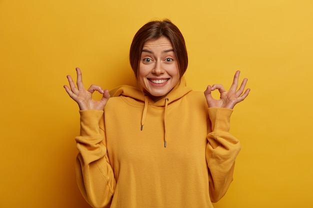 Photo gratuite heureuse jeune femme aux cheveux noirs dit que ça sonne bien, confirme quelque chose, tout est sous contrôle et va bien, approuve la promotion, a une expression joyeuse, est d'accord avec la personne, porte un sweat-shirt jaune.