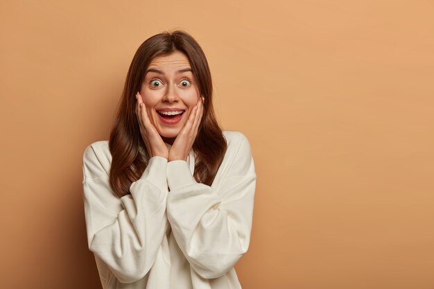 Heureuse jeune femme aux cheveux noirs impressionnée par une réduction de vente folle, garde les paumes sur les joues, porte un sweat-shirt blanc, entend des nouvelles intéressantes