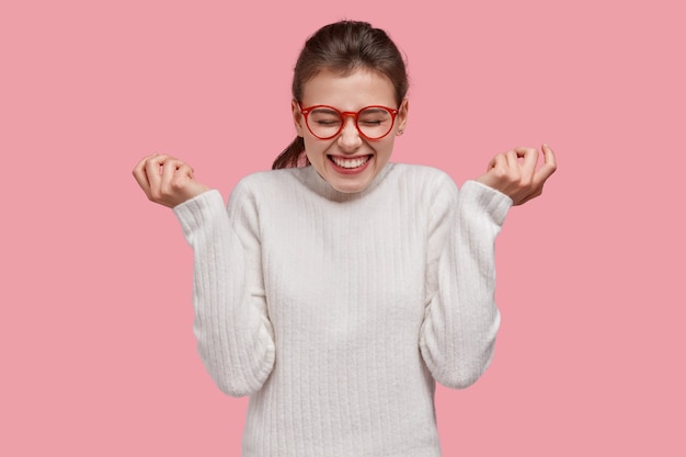 Heureuse jeune femme aux cheveux noirs lève les mains, fait des gestes avec joie, a un large sourire, se sent satisfaite, exprime des émotions positives