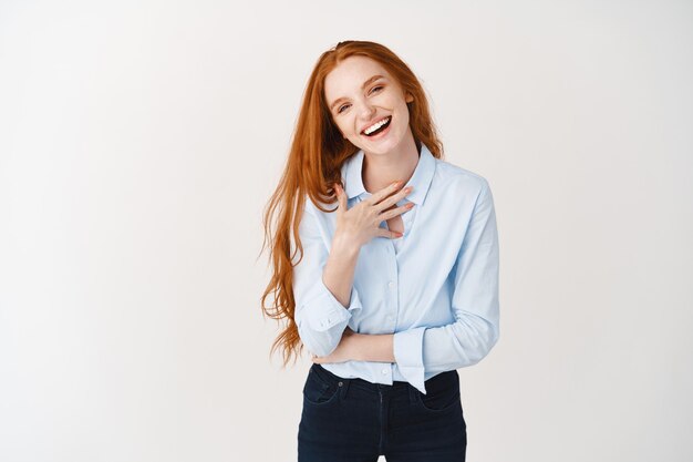 Heureuse jeune femme aux longs cheveux roux pointant sur elle-même, riant et ayant l'air confiante, debout sur un mur blanc