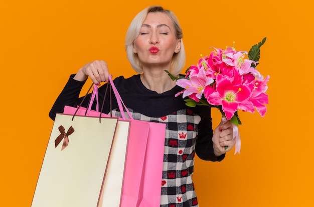 Photo gratuite heureuse jeune femme en belle robe tenant un bouquet de fleurs et de sacs en papier avec des cadeaux soufflant un baiser célébrant la journée internationale de la femme debout sur le mur orange
