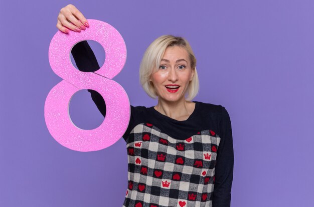 Heureuse jeune femme en belle robe tenant le numéro huit en carton à l'avant souriant joyeusement célébrant la journée internationale de la femme debout sur le mur violet