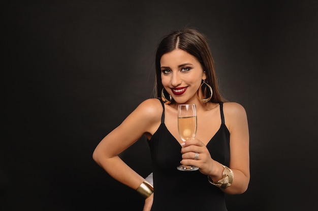 Heureuse jeune femme avec des bijoux d'or en robe noire, boire du champagne